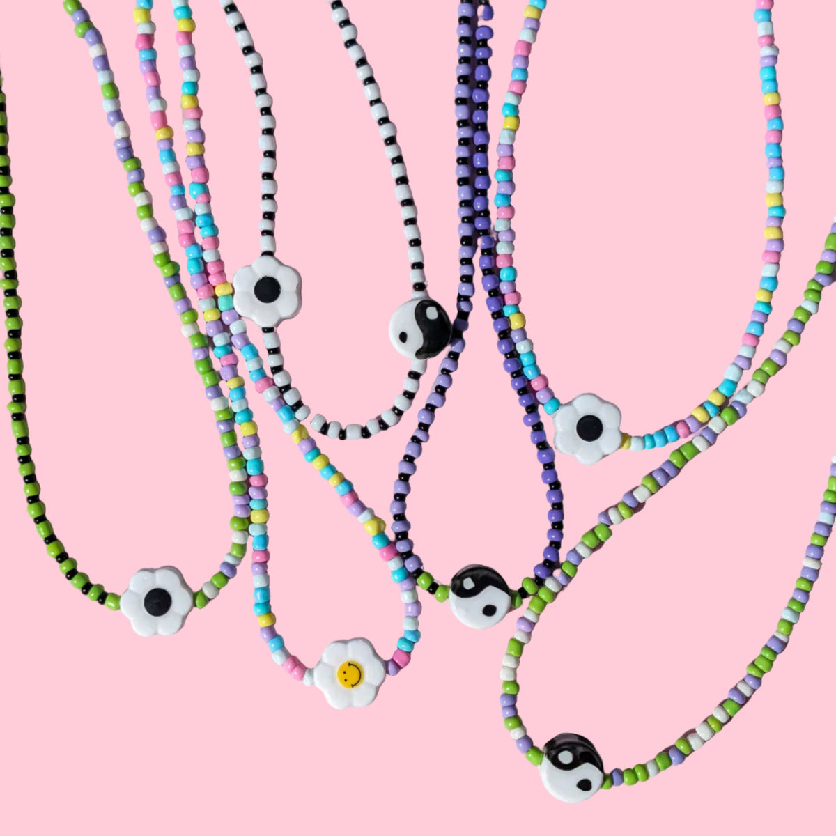 A group of beaded necklaces with white flower charms and black-and-white evil eye beads, displayed on a pink background.