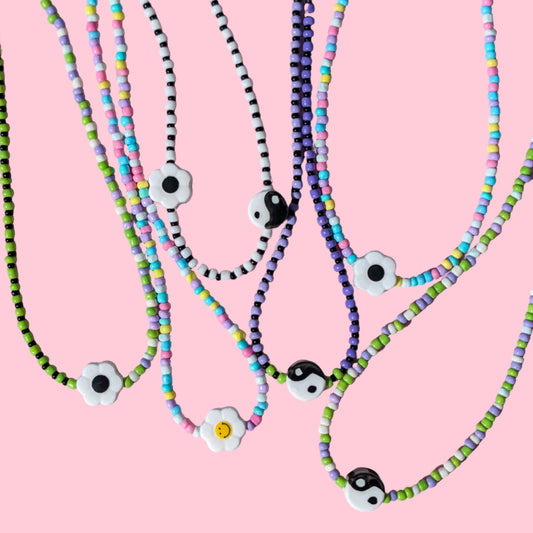 A group of beaded necklaces with white flower charms and black-and-white evil eye beads, displayed on a pink background.