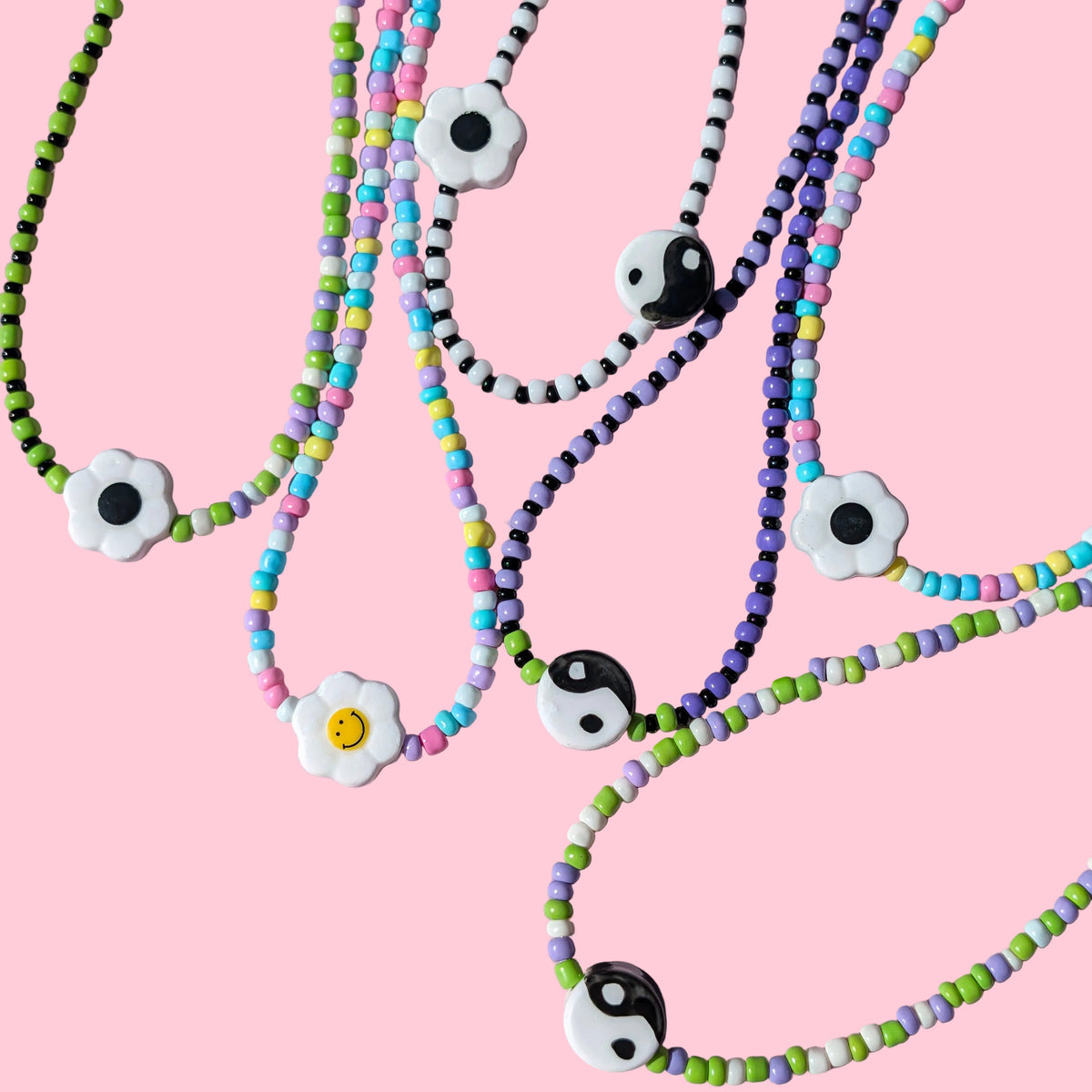 Several beaded necklaces arranged on a pink background, featuring white flower charms with black and yellow centers and black-and-white yin-yang style beads.