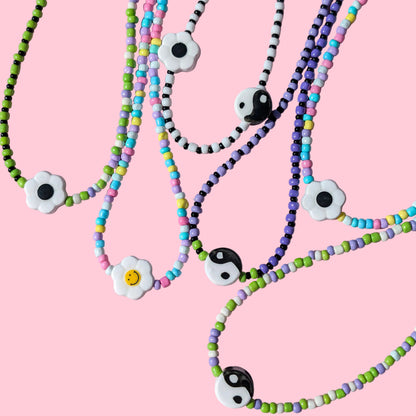 Several beaded necklaces arranged on a pink background, featuring white flower charms with black and yellow centers and black-and-white yin-yang style beads.