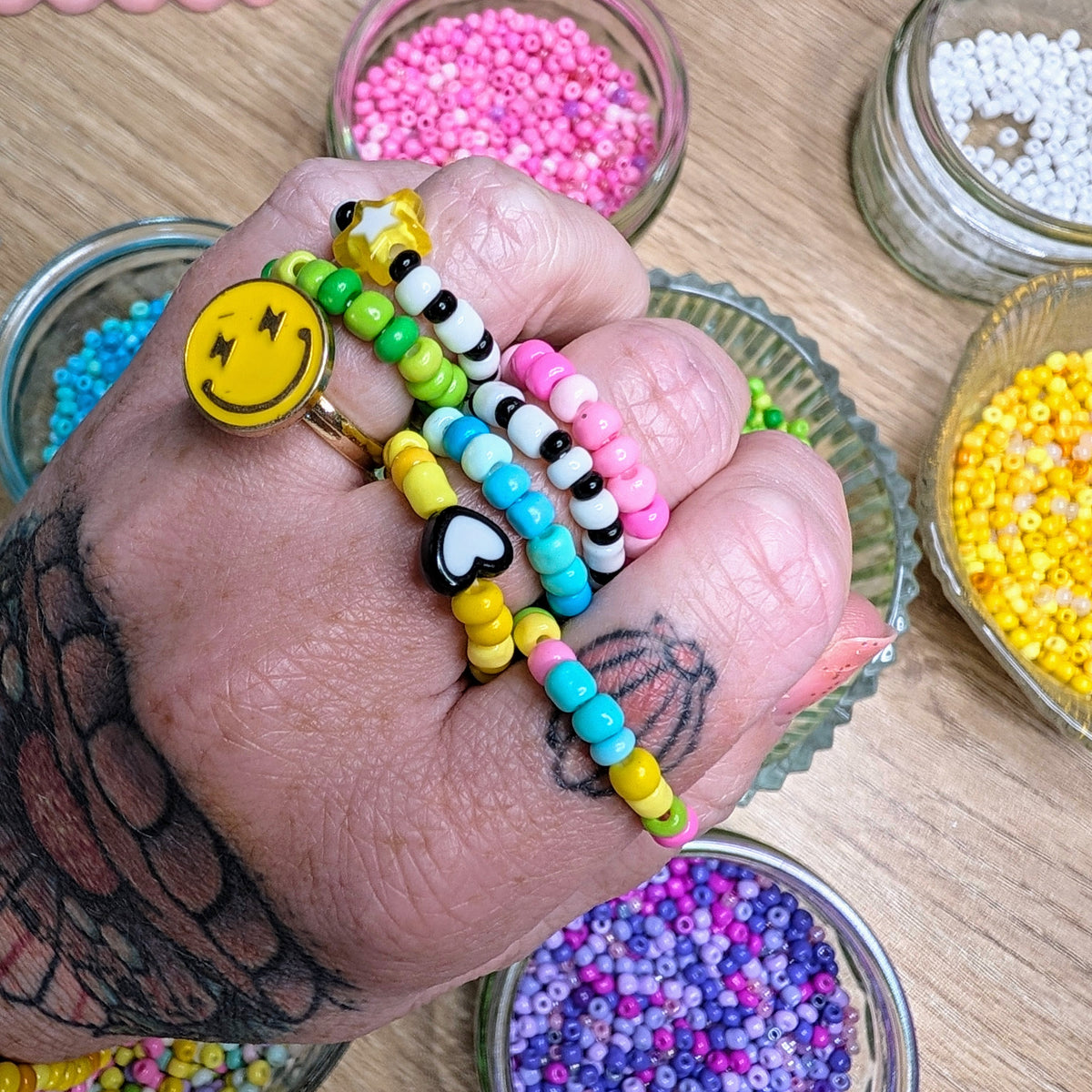 A close-up of a hand wearing several colorful beaded rings, surrounded by small glass bowls filled with multicolored craft beads on a wooden surface.