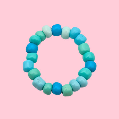 A beaded stretch ring made of faceted cylindrical beads in shades of turquoise, teal, mint green, and light blue, arranged in a circular shape on a plain off-white background.