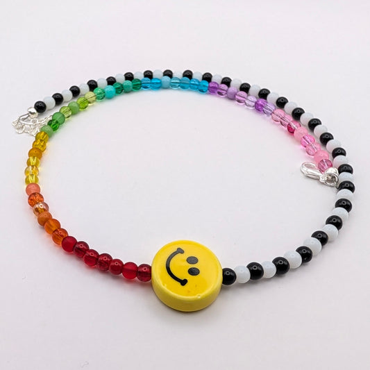 Tilly handmade necklace with rainbow and alternating black and white glass beads, centred with a yellow ceramic smiley face.