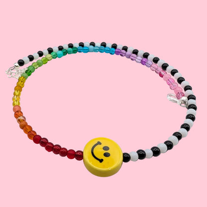 Tilly handmade necklace with rainbow and alternating black and white glass beads, centred with a yellow ceramic smiley face on a pink background.