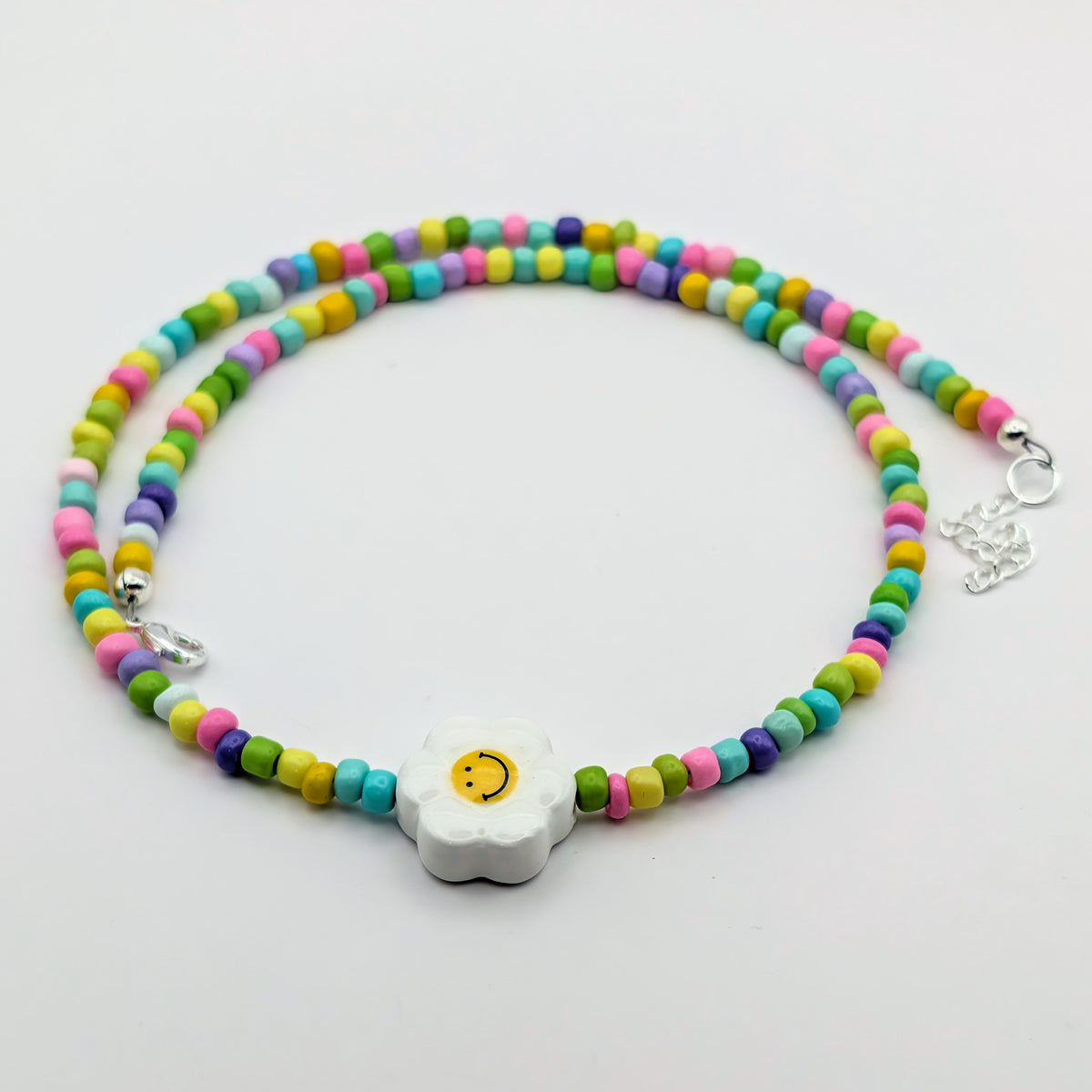 A multicoloured beaded necklace with a central white daisy charm featuring a yellow smiley face. The beads are arranged in two rows of alternating pastel pink, blue, yellow, and purple, with a silver lobster clasp and extender chain.