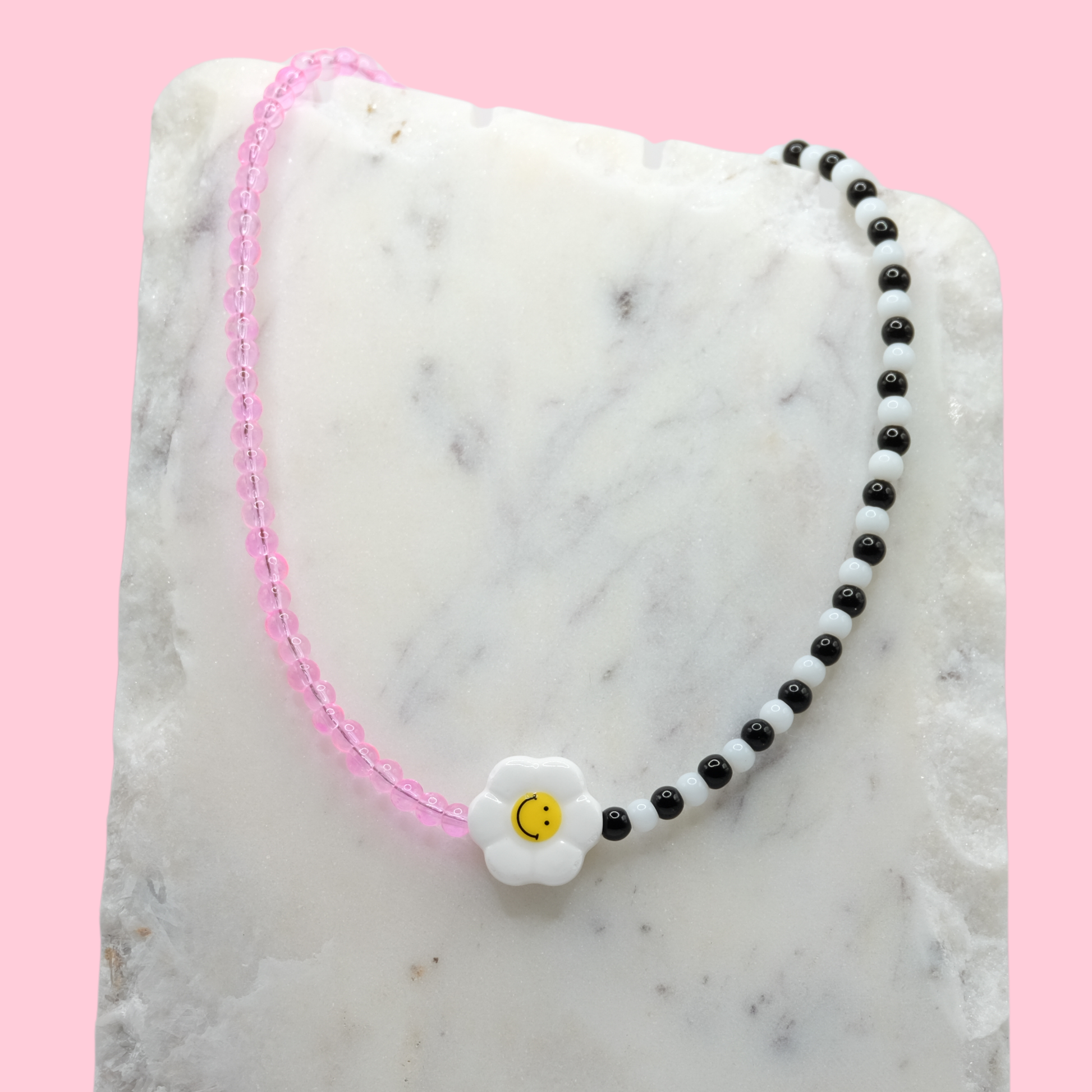 A handmade beaded necklace composed of translucent pink 4mm glass beads and alternating black-and-white glass beads, featuring a ceramic daisy smiley face bead at the centre, displayed on a white marble slab.