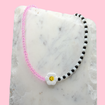 A handmade beaded necklace composed of translucent pink 4mm glass beads and alternating black-and-white glass beads, featuring a ceramic daisy smiley face bead at the centre, displayed on a white marble slab.