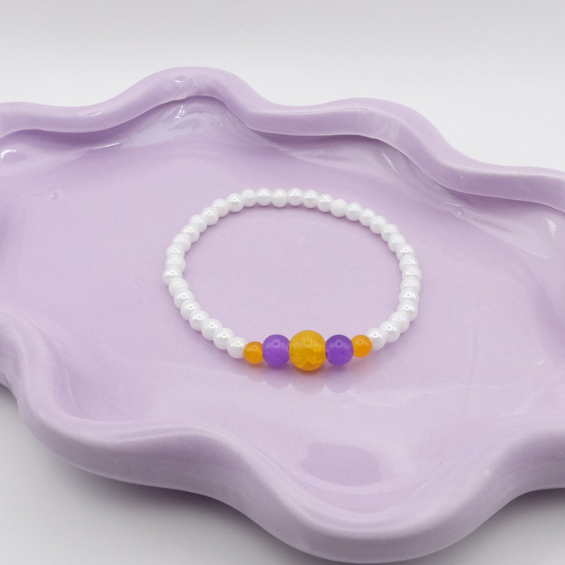 An elastic beaded bracelet made from 4mm metallic white beads, spotlighting five front feature beads—4mm, 6mm, 8mm, 6mm, and 4mm—in bright yellow and dark purple. Photographed on a wavy lavender dish against a clean, light background.
