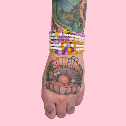 A tattooed arm featuring octopus and seashell art, adorned with a stack of elastic beaded bracelets in varying shades of purple and yellow, posed against a bright pink background