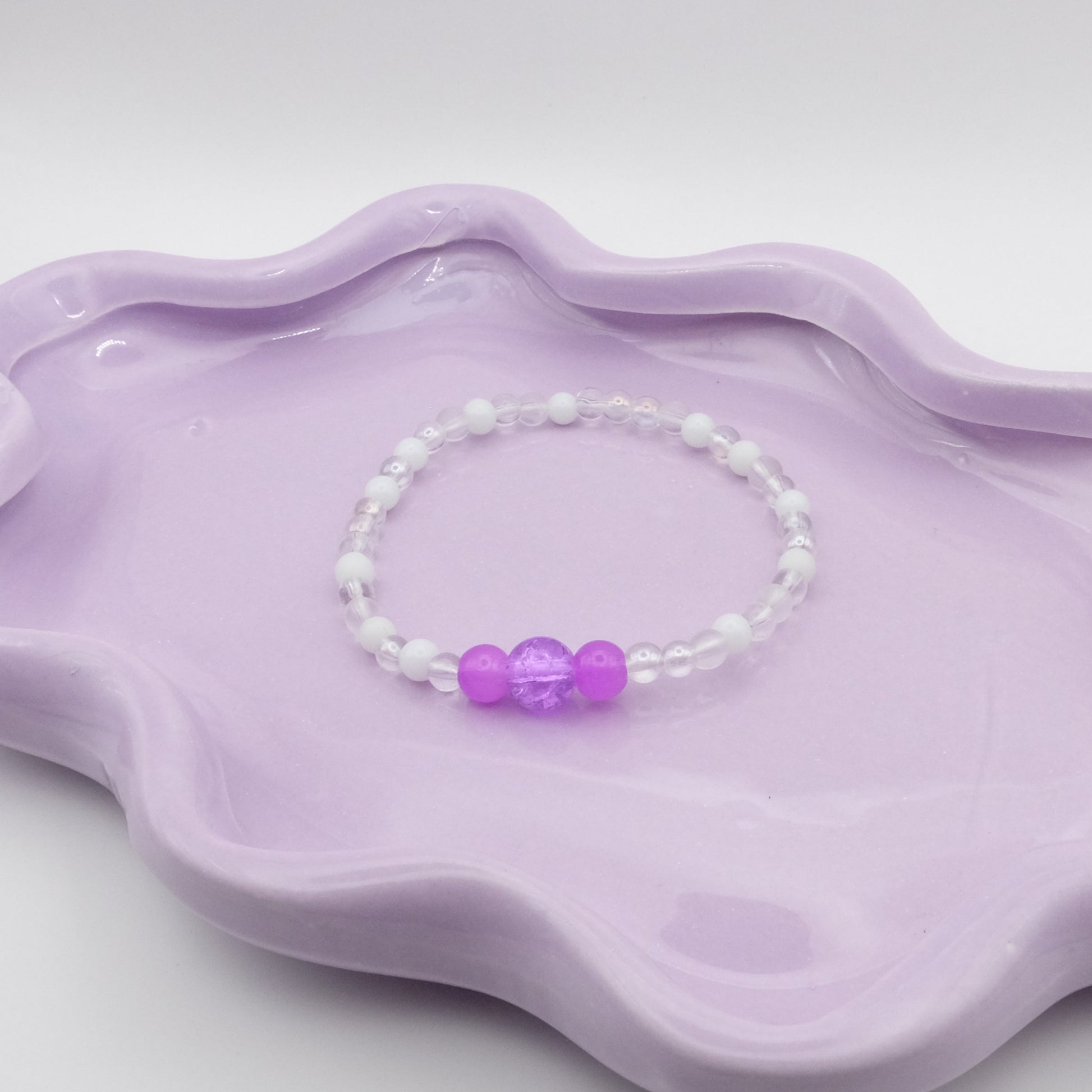 An elastic beaded bracelet made entirely of 4mm translucent and white beads, with a five-bead focal arrangement—4mm, 6mm, 8mm, 6mm, and 4mm—in purple. Resting on a wavy lavender dish, it’s set against a light, minimal backdrop.