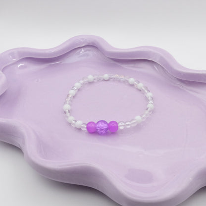 An elastic beaded bracelet made entirely of 4mm translucent and white beads, with a five-bead focal arrangement—4mm, 6mm, 8mm, 6mm, and 4mm—in purple. Resting on a wavy lavender dish, it’s set against a light, minimal backdrop.