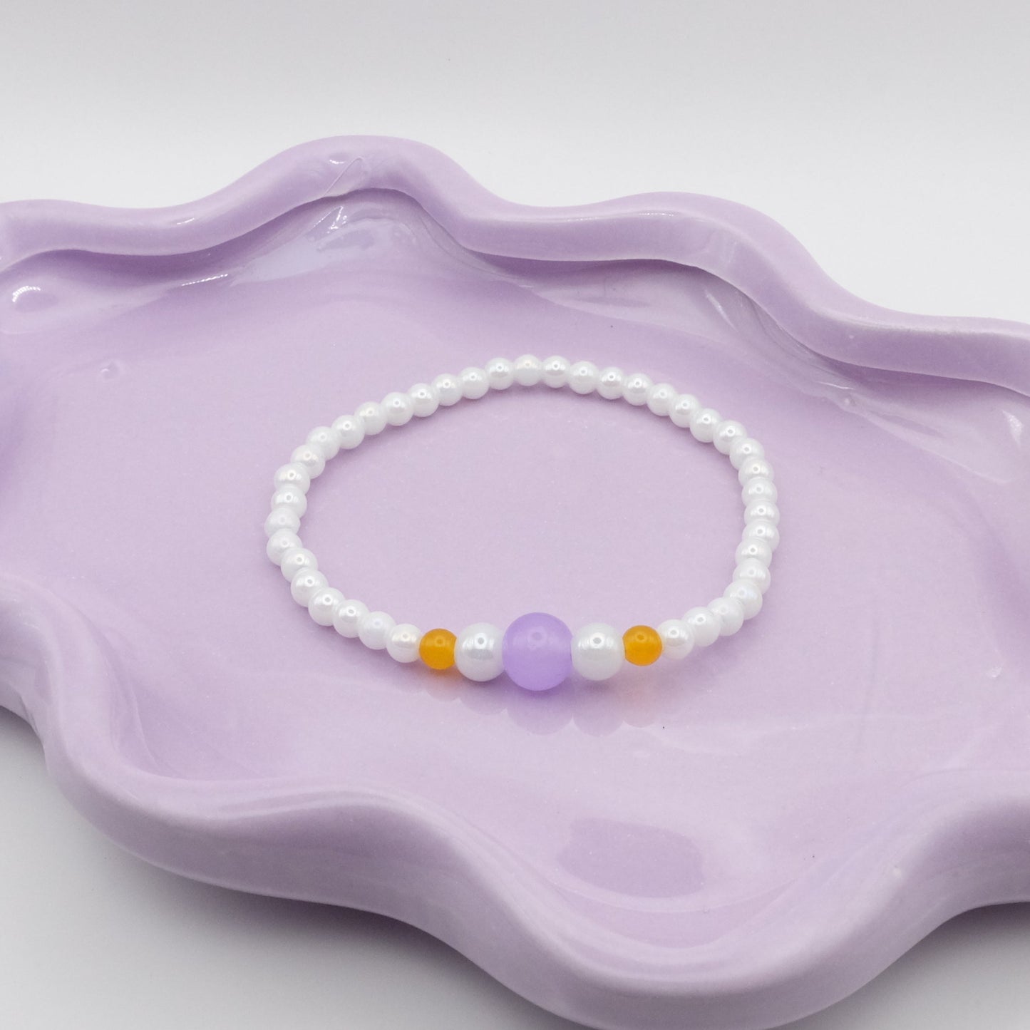 An elastic beaded bracelet made from 4mm metallic white beads, showcasing five front focal beads—4mm, 6mm, 8mm, 6mm, and 4mm—in lavender purple, metallic white, and yellow. Arranged on a wavy lavender dish, it’s set against a gentle, neutral background.