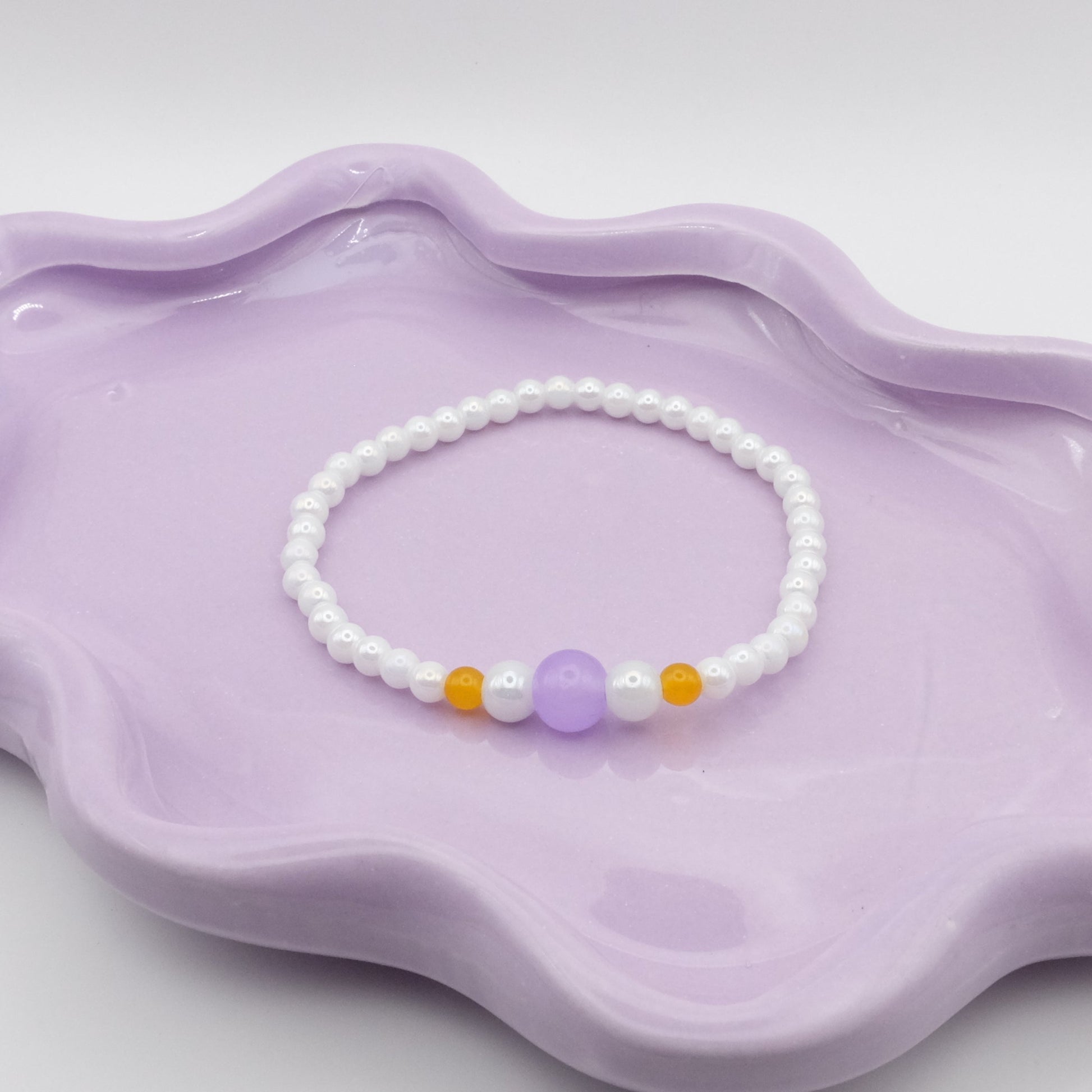 An elastic beaded bracelet made from 4mm metallic white beads, showcasing five front focal beads—4mm, 6mm, 8mm, 6mm, and 4mm—in lavender purple, metallic white, and yellow. Arranged on a wavy lavender dish, it’s set against a gentle, neutral background.