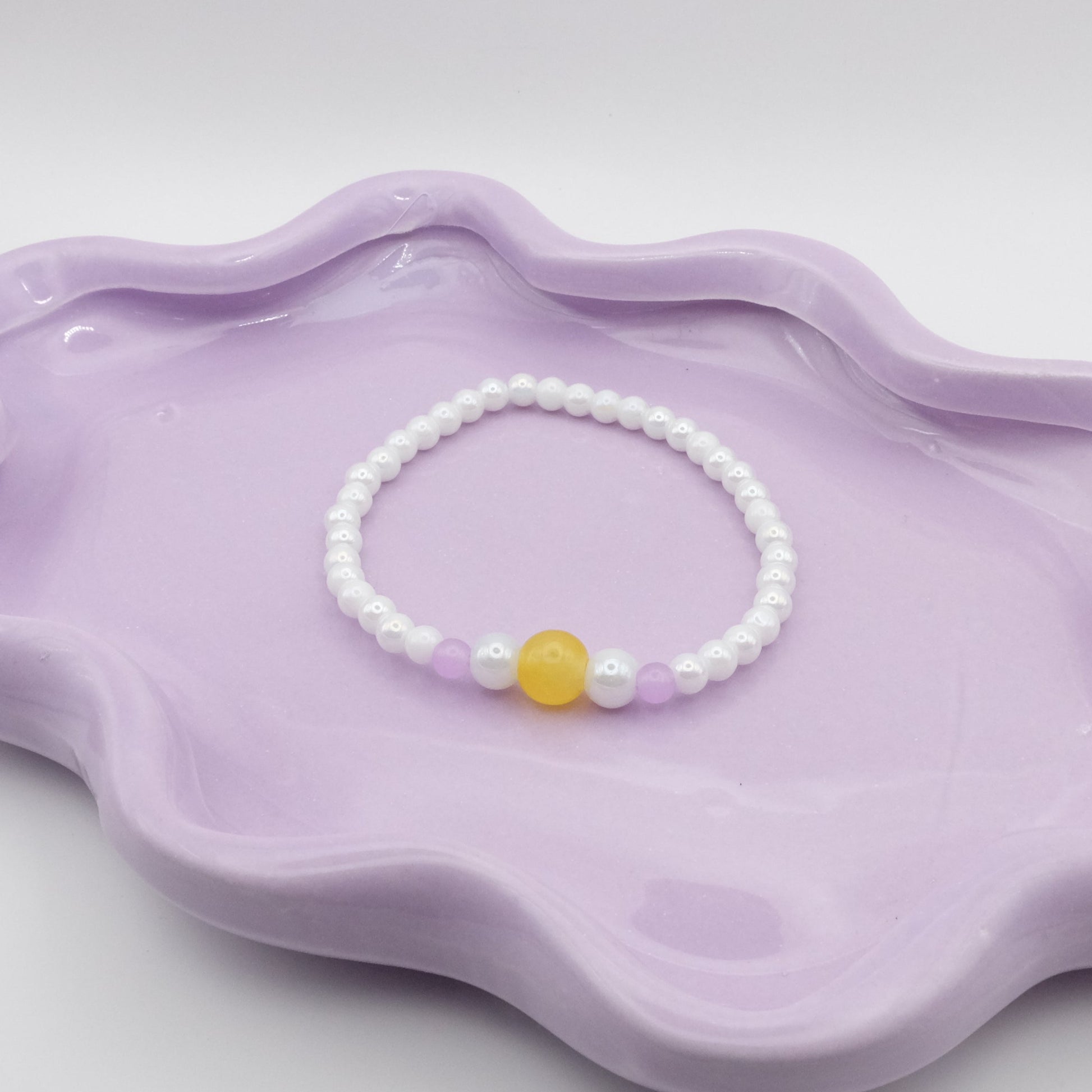 An elastic beaded bracelet composed of 4mm metallic white beads, showcasing a five-bead focal sequence—4mm, 6mm, 8mm, 6mm, and 4mm—in yellow, metallic white, and lavender purple. Photographed on a wavy lavender dish with a soft, neutral background.