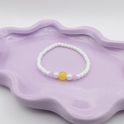 An elastic beaded bracelet composed of 4mm metallic white beads, showcasing a five-bead focal sequence—4mm, 6mm, 8mm, 6mm, and 4mm—in yellow, metallic white, and lavender purple. Photographed on a wavy lavender dish with a soft, neutral background.