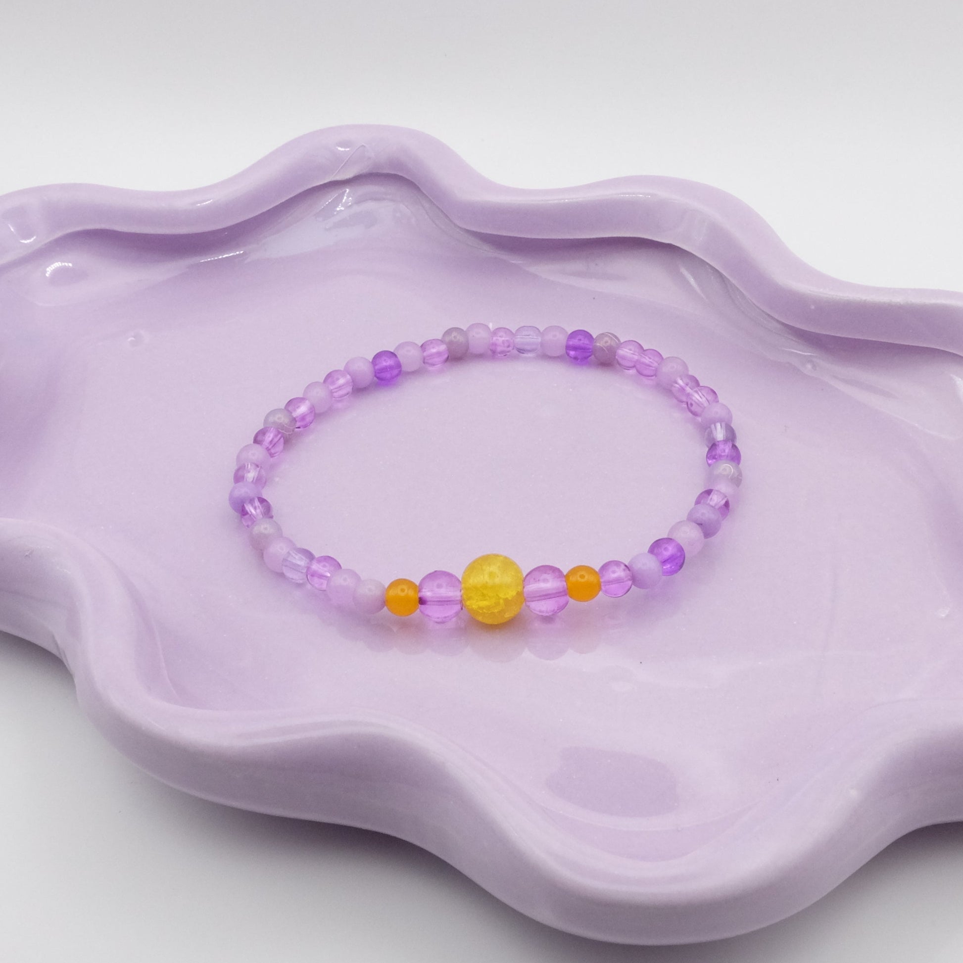 An elastic beaded bracelet featuring 4mm purple beads with a five-bead focal arrangement—4mm, 6mm, 8mm, 6mm, and 4mm—in yellow and purple. Displayed on a wavy lavender dish with a softly lit backdrop.