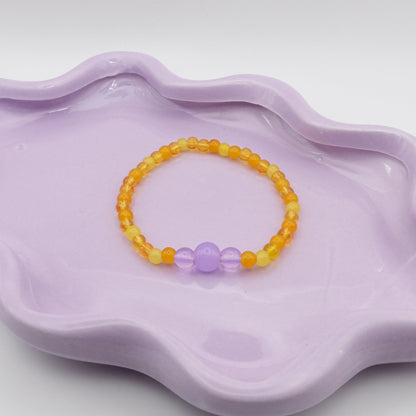 An elastic beaded bracelet crafted entirely from 4mm glass beads in a mix of yellows highlighted by a feature of 4mm, 6mm, and 8mm glass beads  highlighted by lavender purple focal beads, displayed on a wavy lavender dish against a simple, light backdrop.
