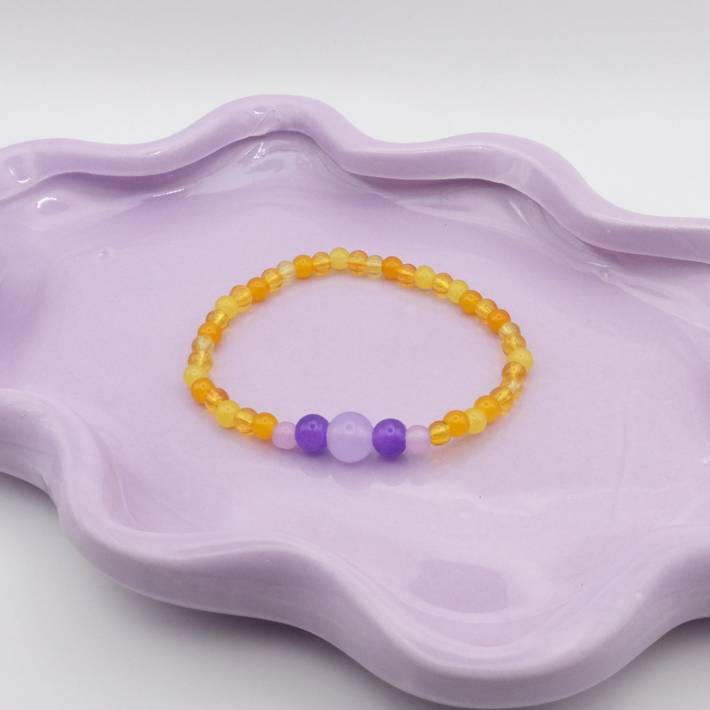 An elastic beaded bracelet created with 4mm yellow beads and a whimsical five-bead focal sequence—4mm, 6mm, 8mm, 6mm, and 4mm—featuring lavender purple and dark purple accents. Placed on a wavy lavender dish, it’s displayed in front of a simple, neutral background.