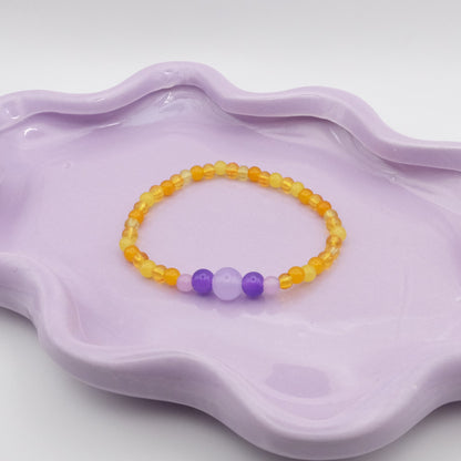 An elastic beaded bracelet created with 4mm yellow beads and a whimsical five-bead focal sequence—4mm, 6mm, 8mm, 6mm, and 4mm—featuring lavender purple and dark purple accents. Placed on a wavy lavender dish, it’s displayed in front of a simple, neutral background.