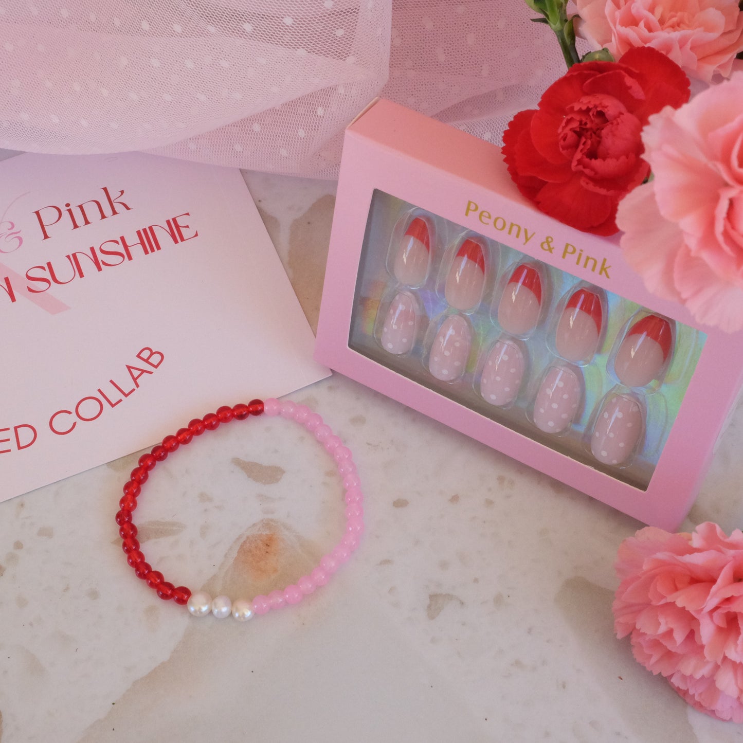 Pink and red beaded bracelet with a box of false nails and flowers on a marble surface.
