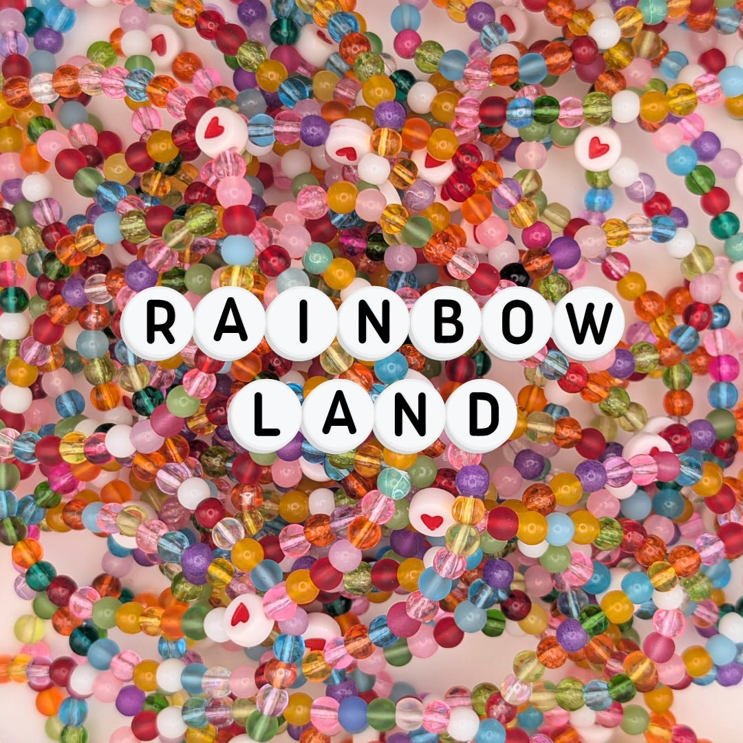 A vibrant pile of elastic beaded bracelets made from colourful 4mm glass beads, with heart accents and letter beads spelling “RAINBOWLAND.” A joyful handmade jewellery collection perfect for dopamine dressing, quirky jewellery, and colourful accessories.