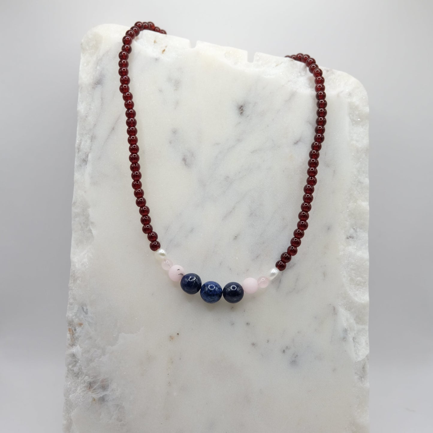 A beaded necklace composed of deep red glass beads - featuring lapis gemstone beads, pink opal accents and freshwater pearl nuggets at the centre, shown on a white marble background.