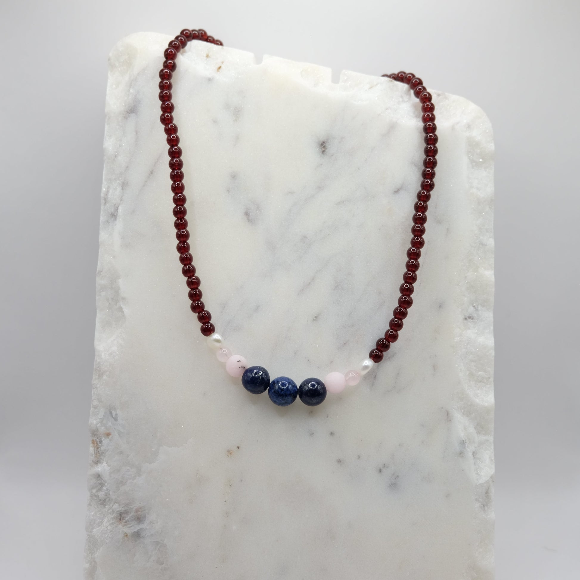 A beaded necklace composed of deep red glass beads - featuring lapis gemstone beads, pink opal accents and freshwater pearl nuggets at the centre, shown on a white marble background.