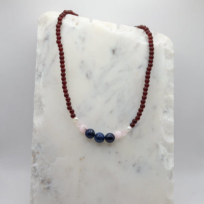 A beaded necklace composed of deep red glass beads - featuring lapis gemstone beads, pink opal accents and freshwater pearl nuggets at the centre, shown on a white marble background.
