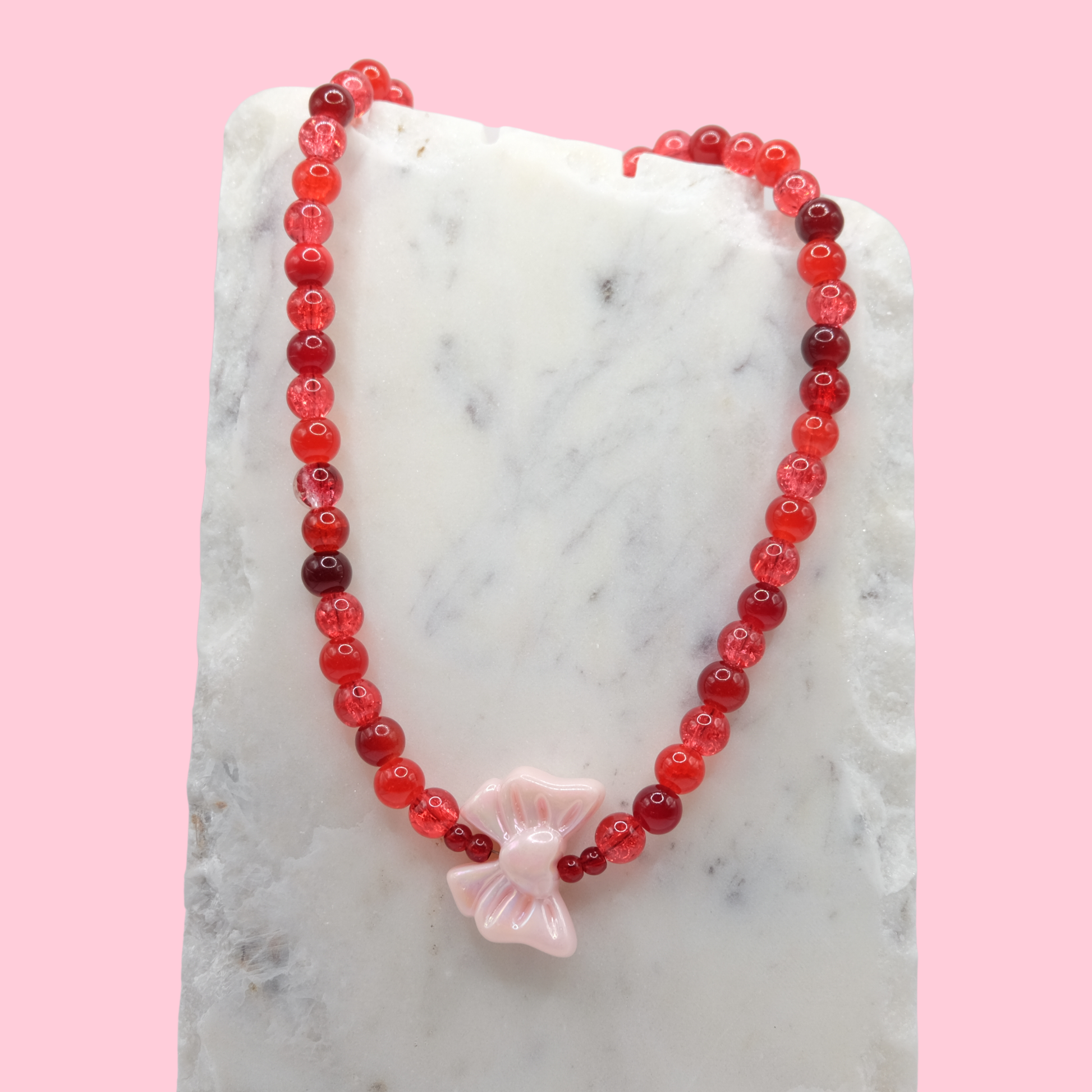 Necklace with an assortment of red glass beads and an oversized pink acrylic bow charm on a marble surface with a pink background.