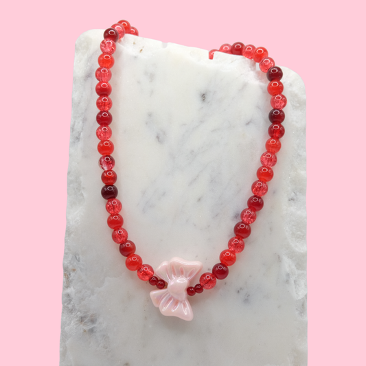 Necklace with an assortment of red glass beads and an oversized pink acrylic bow charm on a marble surface with a pink background.