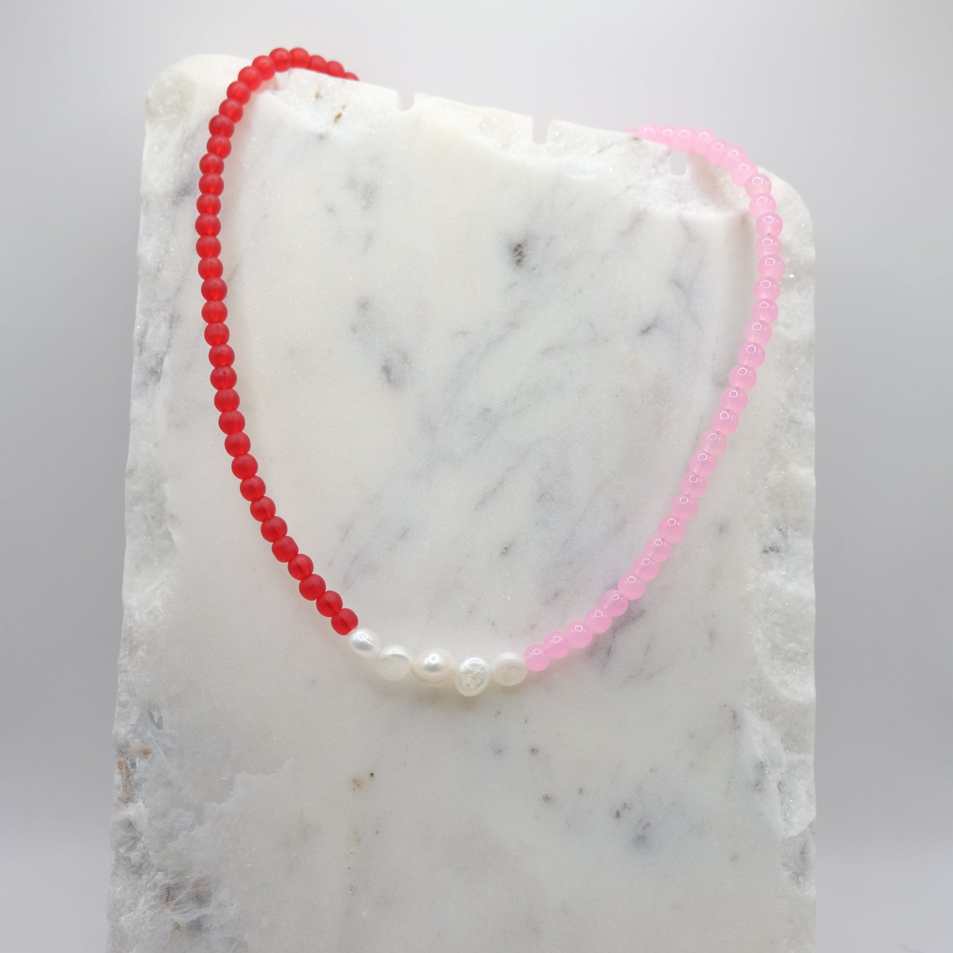A beaded necklace composed of matte red and pink glass beads - featuring a centre row of freshwater pearl nuggets, shown on a white marble background.