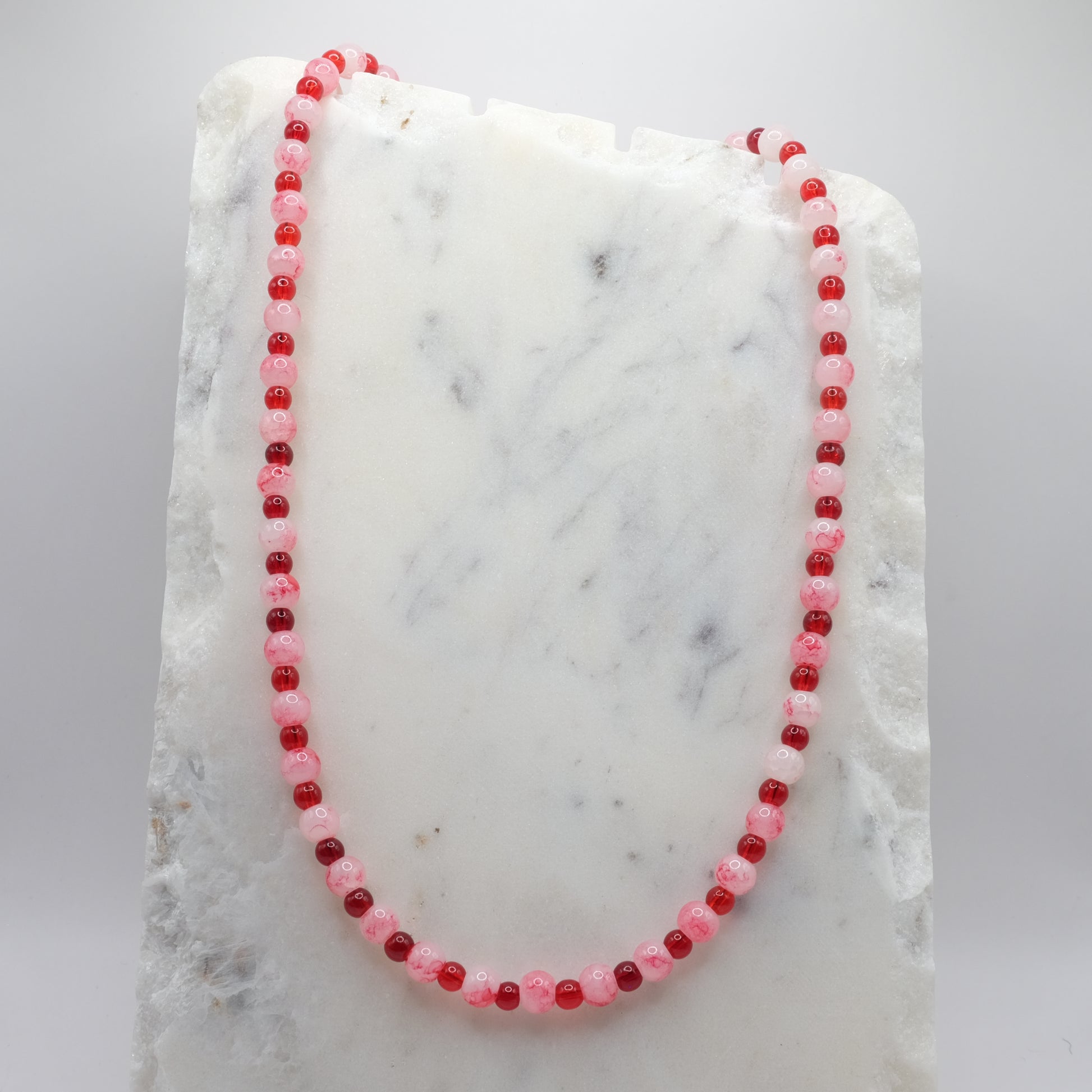 A beaded necklace composed of alternating 4mm and 6mm cherry red and marbled white and red glass beads - featuring glossy finishes, shown on a white marble background.