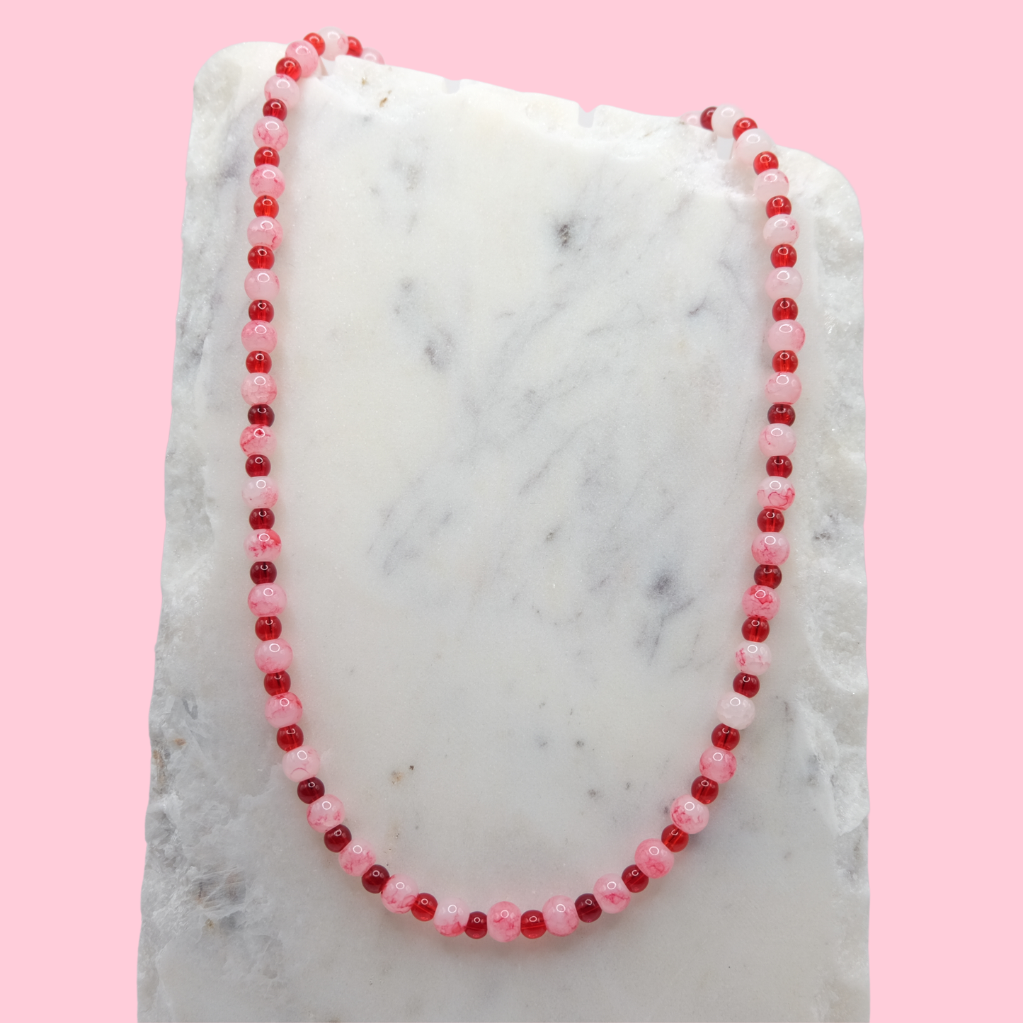 A beaded necklace composed of alternating 4mm and 6mm cherry red and marbled white and red glass beads - featuring glossy finishes, shown on a white marble background. Background is pink