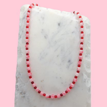 A beaded necklace composed of alternating 4mm and 6mm cherry red and marbled white and red glass beads - featuring glossy finishes, shown on a white marble background. Background is pink