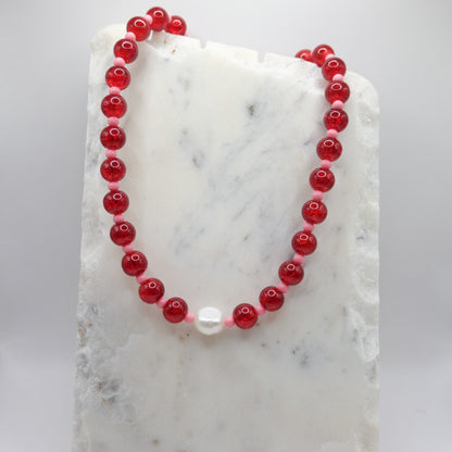 A beaded necklace composed of red crackle glass beads and small pink beads - featuring a central freshwater pearl, shown on a white marble background.