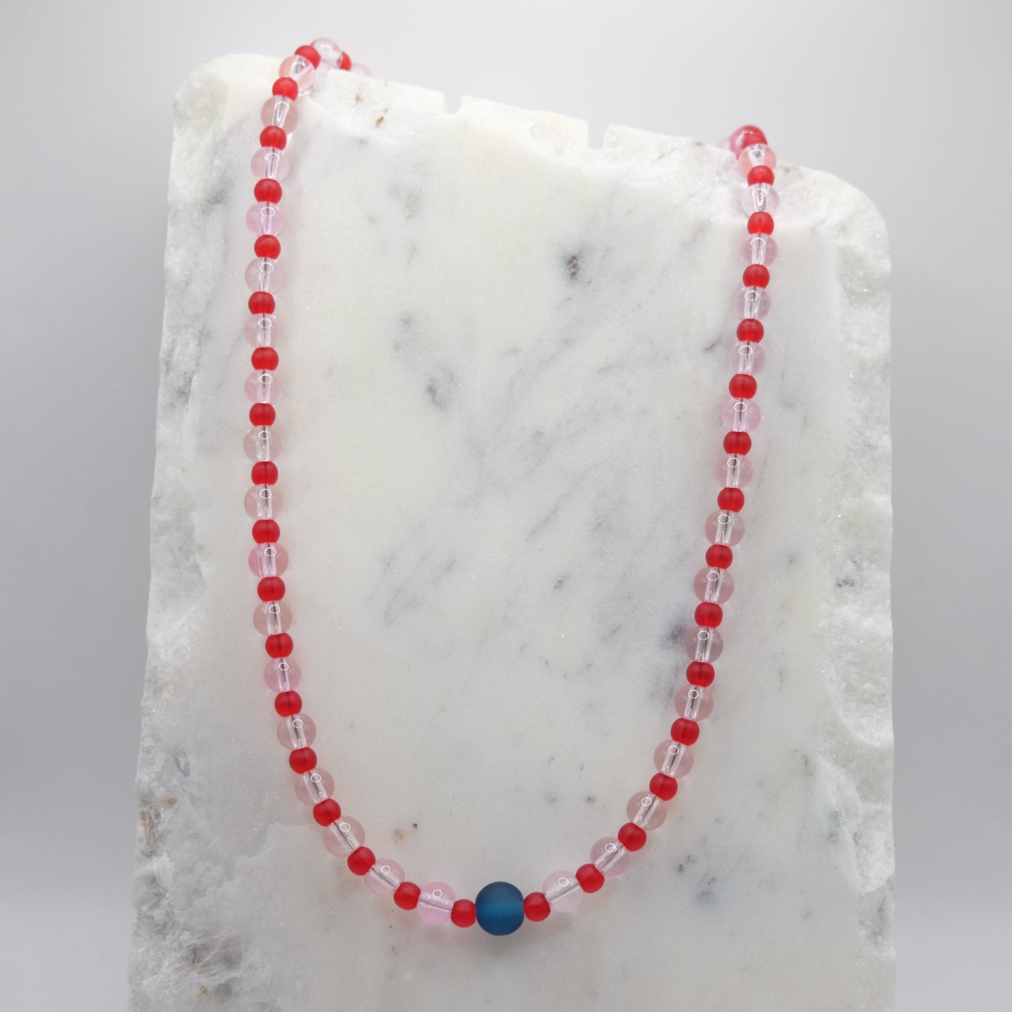 A beaded necklace composed of 6mm pale pink clear glass beads and 4mm matte red beads - featuring a single matte navy blue centre bead, shown on a white marble background.