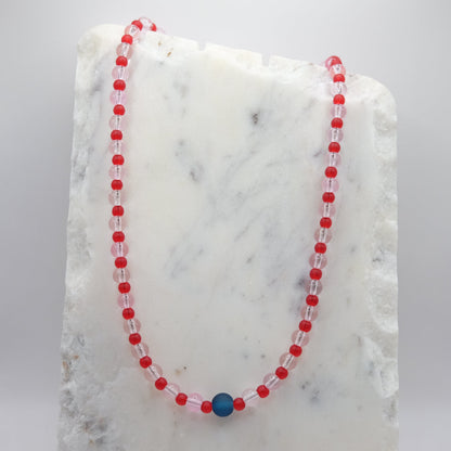 A beaded necklace composed of 6mm pale pink clear glass beads and 4mm matte red beads - featuring a single matte navy blue centre bead, shown on a white marble background.