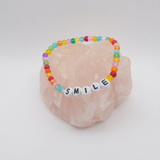 An elastic beaded bracelet composed of vibrant 4mm glass beads in red, orange, yellow, green, aqua, blue, purple and pink with white acrylic letter beads spelling “SMILE,” displayed on a pale pink crystal rock.