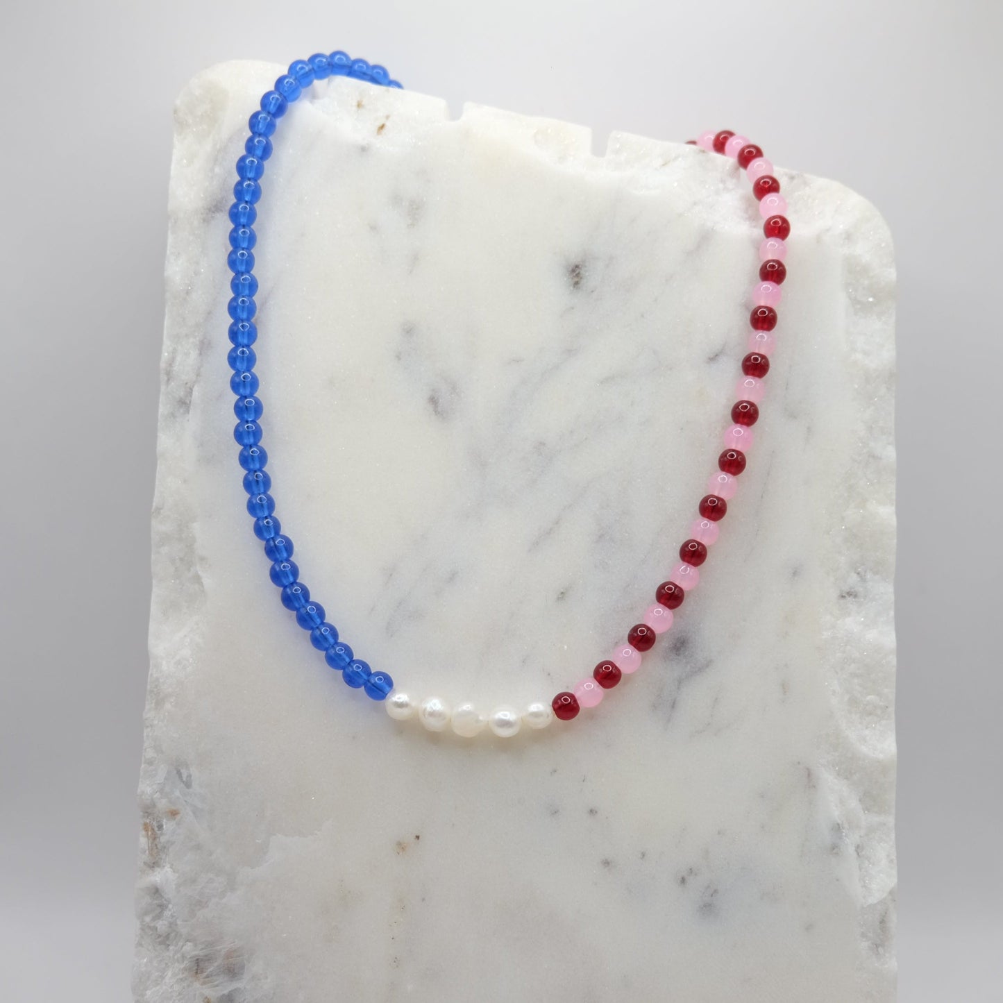 A beaded necklace composed of red, pink and blue glass beads—featuring a centre row of freshwater pearl nuggets, shown on a white marble background.