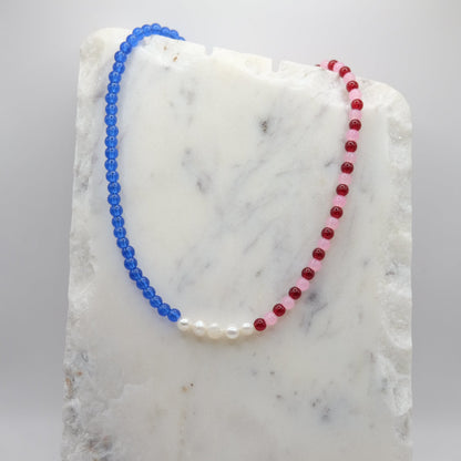 A beaded necklace composed of red, pink and blue glass beads—featuring a centre row of freshwater pearl nuggets, shown on a white marble background.