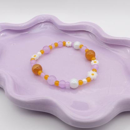 An elastic beaded bracelet bursting with glass beads in purple, yellow, and white hues, including small daisy flowers and ranging from 4mm up to a bold 10mm. It’s displayed on a wavy lavender dish against a light background.