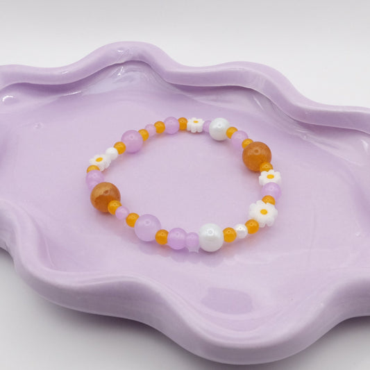 An elastic beaded bracelet bursting with glass beads in purple, yellow, and white hues, including small daisy flowers and ranging from 4mm up to a bold 10mm. It’s displayed on a wavy lavender dish against a light background.