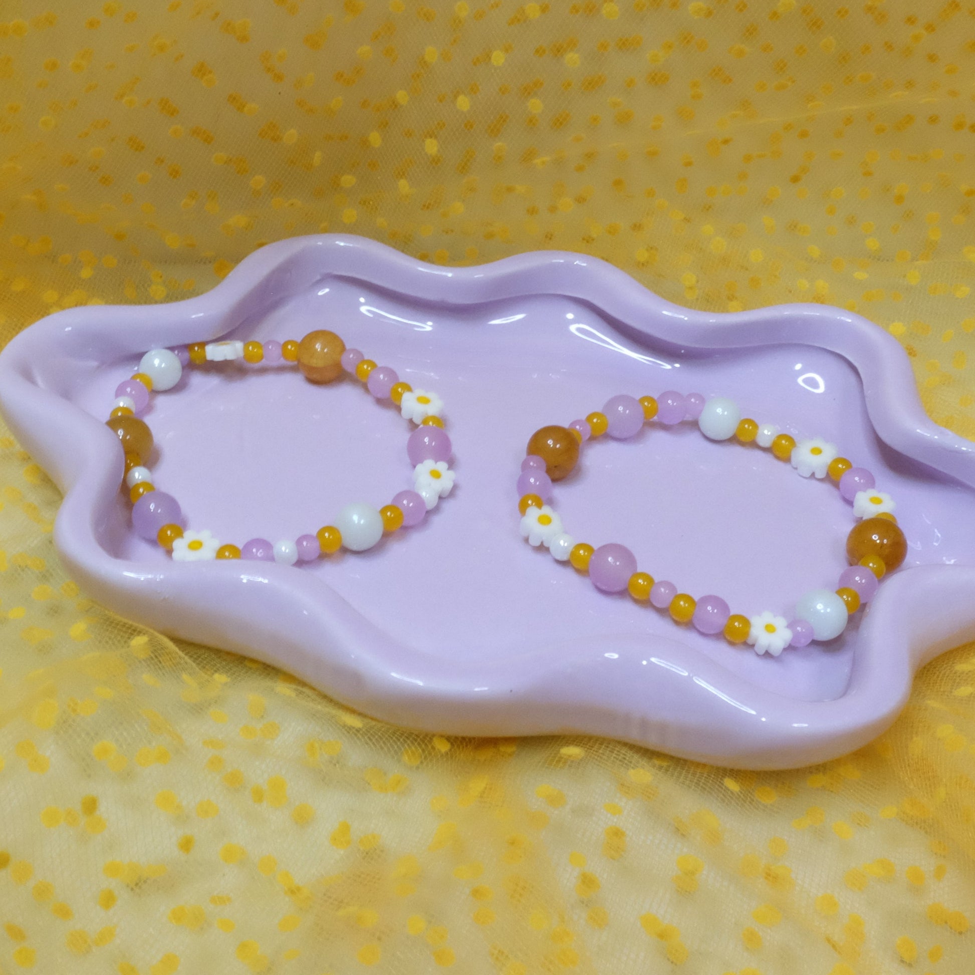Two elastic beaded bracelets in Daisy Pop colorway—each a unique mix of purples, yellows, and crisp white glass beads ranging from 4mm to 10mm, dotted with cheerful daisy beads—are shown side by side on a wavy lavender dish, set against a sunny yellow polka-dot tulle backdrop.