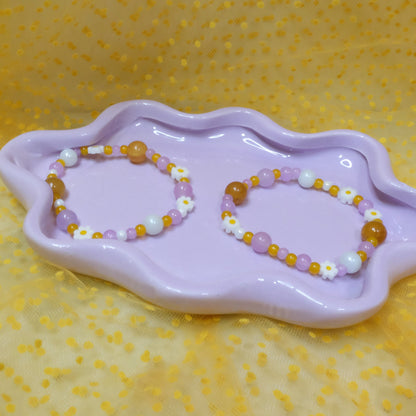 Two elastic beaded bracelets in Daisy Pop colorway—each a unique mix of purples, yellows, and crisp white glass beads ranging from 4mm to 10mm, dotted with cheerful daisy beads—are shown side by side on a wavy lavender dish, set against a sunny yellow polka-dot tulle backdrop.