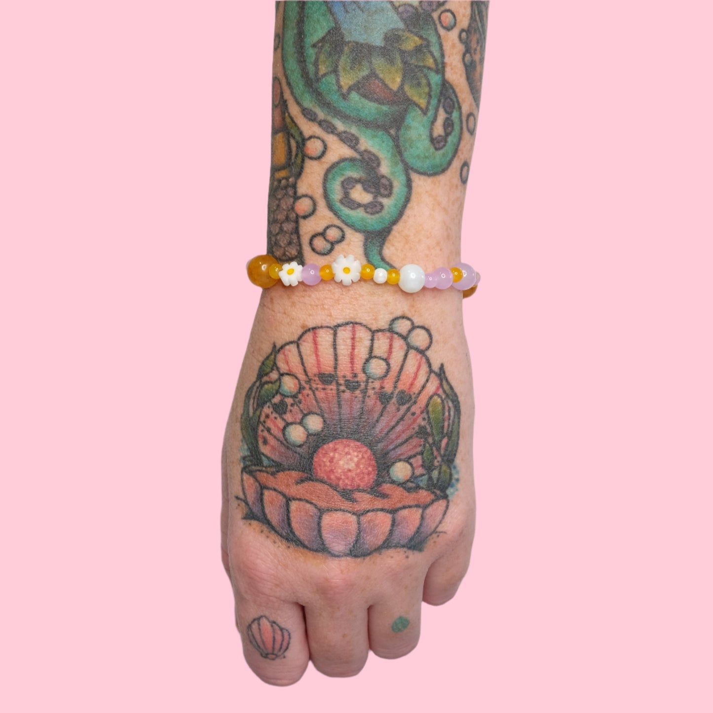 A tattooed arm with octopus and seashell tattoos, wearing an elastic beaded bracelet in yellow and lavender hues accented by small white daisy beads, set against a solid pink background.