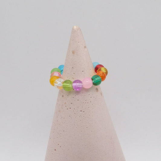 An elastic beaded ring composed of colourful 4mm glass beads in red,  orange, yellow, green, aqua, blue,  purple and pink, displayed on a pale pink ring holder.