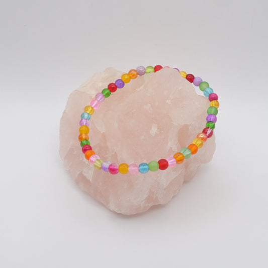 An elastic beaded bracelet composed of colourful 4mm glass beads in red,  orange, yellow, green, aqua, blue,  purple and pink, displayed on a pale pink crystal rock.