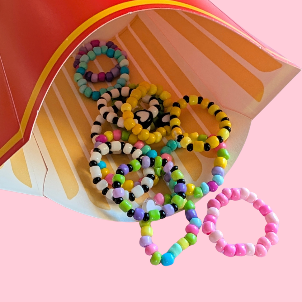 A group of colorful beaded stretch rings spilling out of a red and yellow french fries carton against a solid pink background.