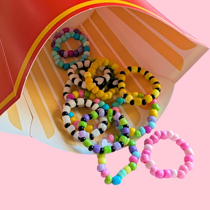 A group of colorful beaded stretch rings spilling out of a red and yellow french fries carton against a solid pink background.