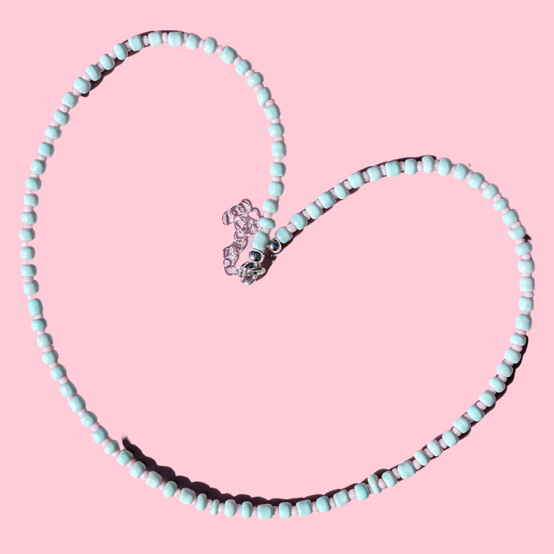 A heart-shaped beaded necklace made of light blue-green beads with a silver-toned lobster clasp, displayed on a solid pink background.